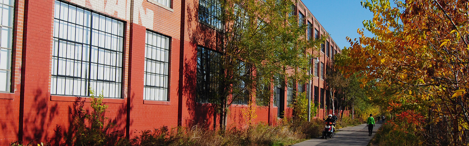 Paved trail runs along the side of a 3-storey industrial building. Bushes and other greenery line the other side of the trail. People can be seen walking along the path.
