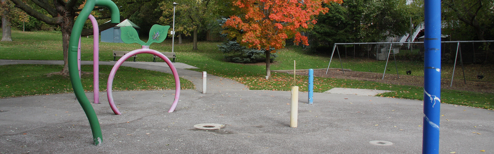 Child-like pipes stick out of the ground around a dry splashpad. A swingset sits beyond it to the right and a paved park trail leads away from it towards two park benches and a green park lawn. Fall trees surround the area.