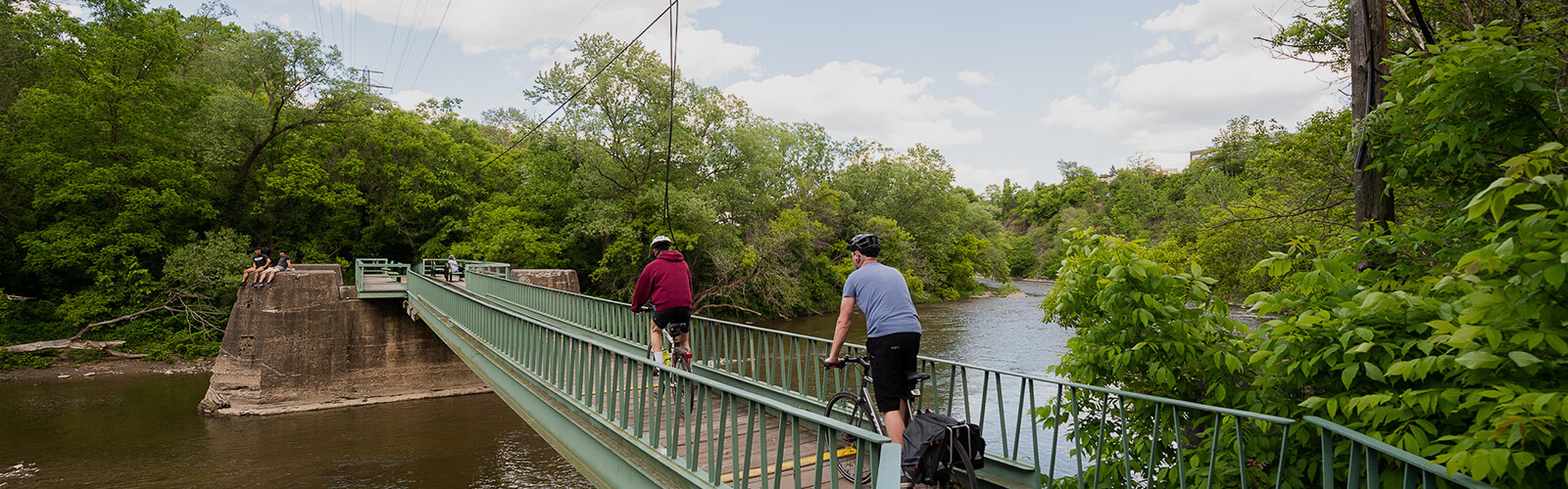 Leading from the foreground, a metal pedestrian bridge crossing the Humbber River. Two cyclists pedal across the bridge. Either side of the river is lined with lush trees and greenery.