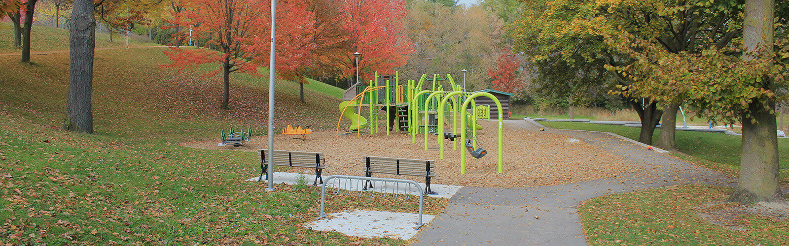 Paved path leading from the foreground to a playground. Fall trees surround the park, along with a couple of park benches and a bike rack.