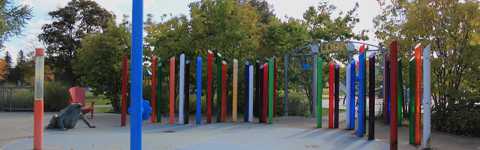 Colourful pillars surround a dry blue circular splash pad. To the left is a large sculpture of a frog, and behind that a red Muskoka chair. Behind the colourful pillars is a line of tall lush trees.