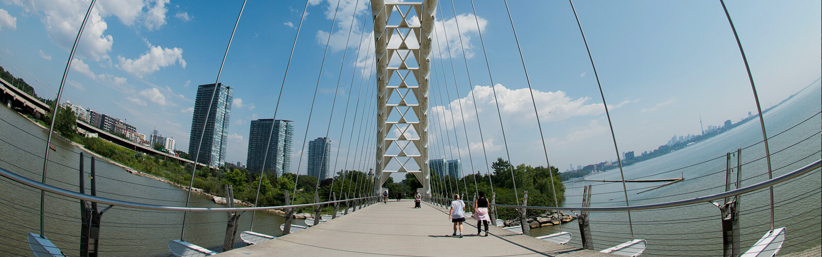 Sunny fish eye photo of the Humber Bay Arch Bridge and people crossing it. On the right side is Lake Ontario and in the distance, the Toronto skyline. On the left, the Humber River and three condo buildings in the background.