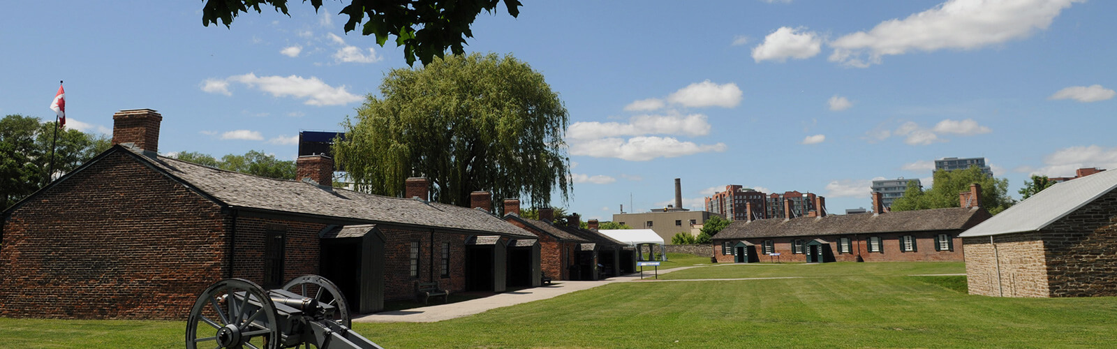 Taken from underneath a lush maple tree, of which the leaves are silhouetted against a clear blue sky. The view is of Fort York, and the historic buildings that are on the grounds.
