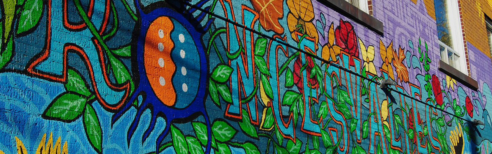 A bright and colourful mural painted on the side of a building with a few windows at the top. The mural features flowers and other greenery and the name "Roncesvalles" painted in large lettering. Painted vines run through the lettering.