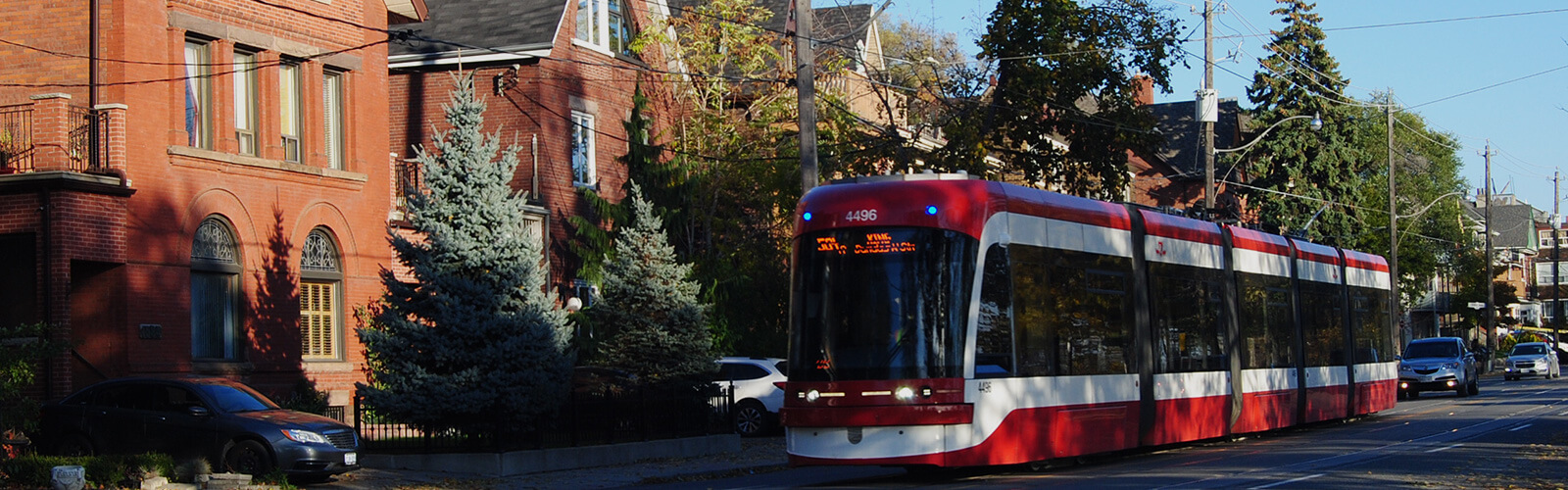 A TTC King Street Streetcar drives along, past several homes with a couple of cars driving behind it.