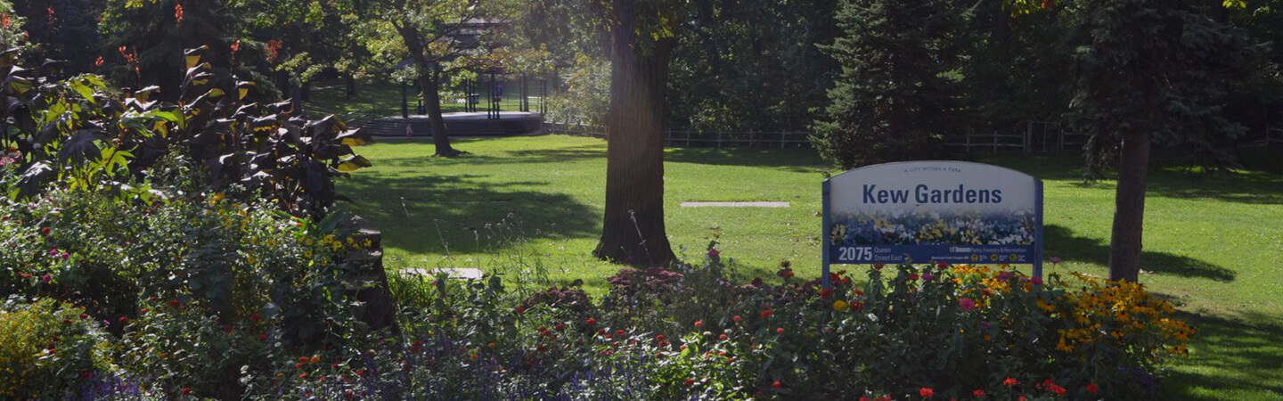 A lush flower bed sits in the foreground with a Toronto Parks sign that reads Kew Gardens. Manicured lawn with several large trees and a large dark gazebo are in the background.