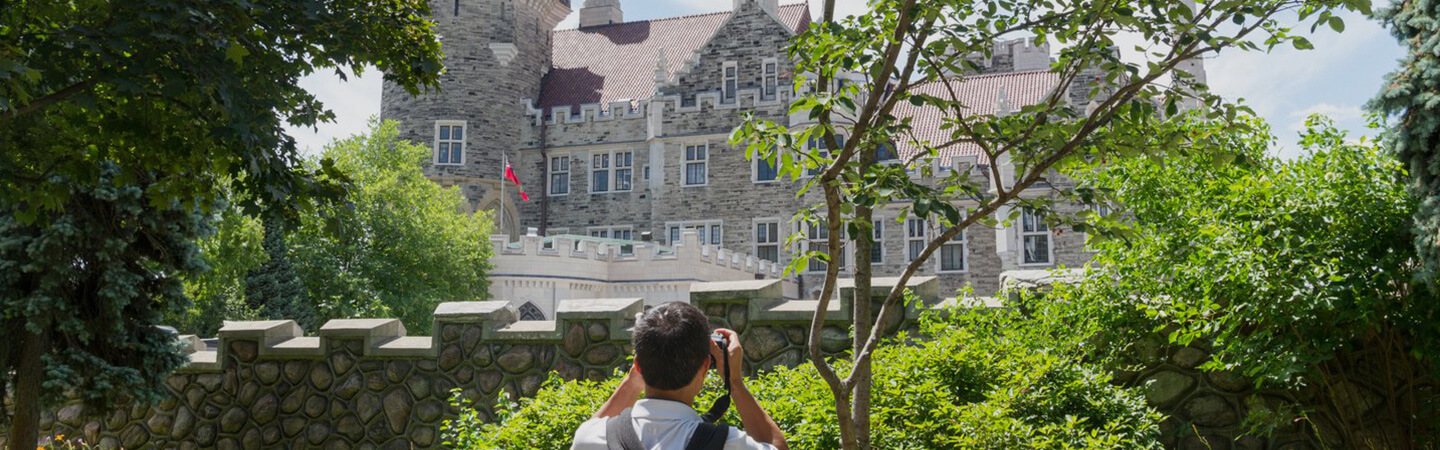 In the foreground, a man in a white t-shirt and black backpack facing away, taking a photo of Casa Loma in the background.