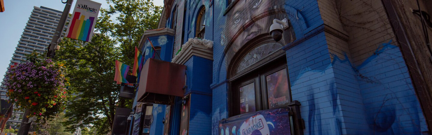 Front of old brick building painted with a large blue mural, and Pride flags hanging off it. In front of the building, a lamp post with hanging flower pots. In the background, a large lush tree and a high-rise condo building.