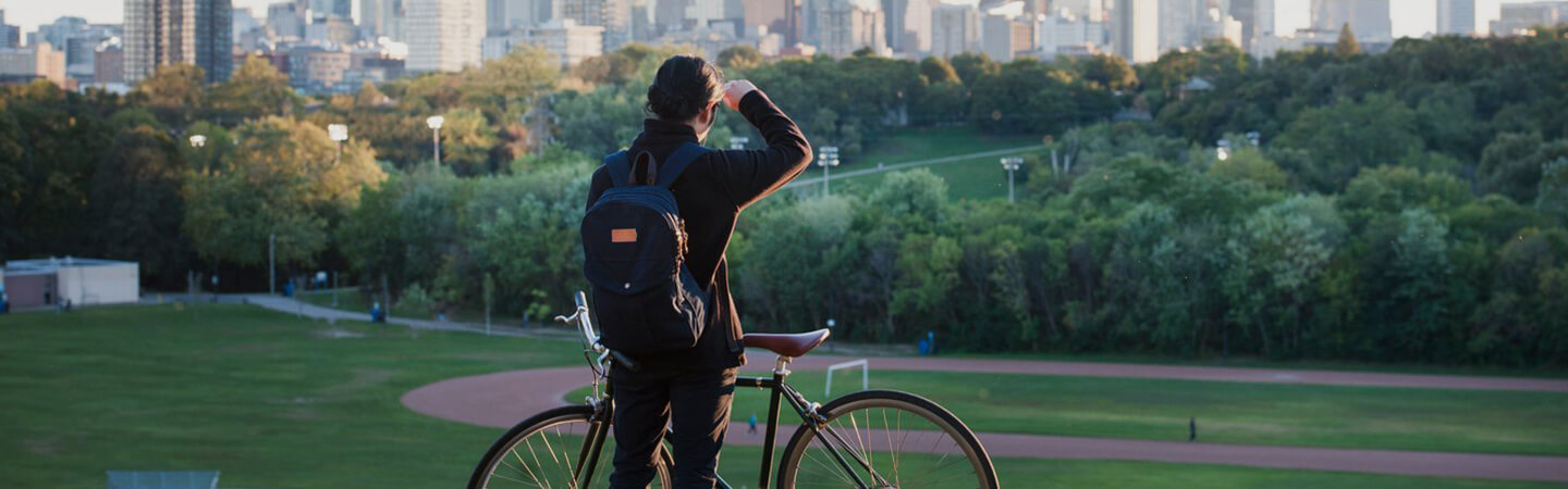 In the foreground, a person stands behind their bicycle facing Riverdale Park and the Don Valley. Lush trees and Toronto's downtown core line the background.