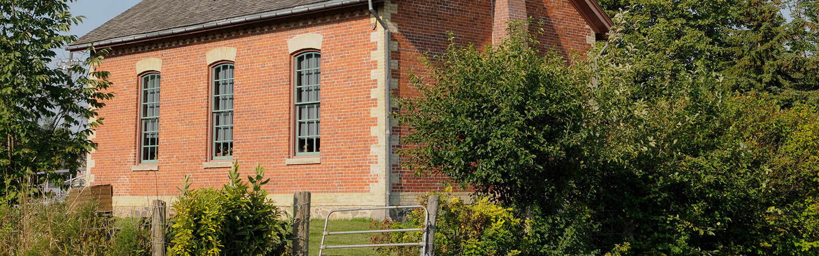 The back of the Zion Schoolhouse surrounded by lush greenery and a well maintained lawn with a wire fence is in the foreground.