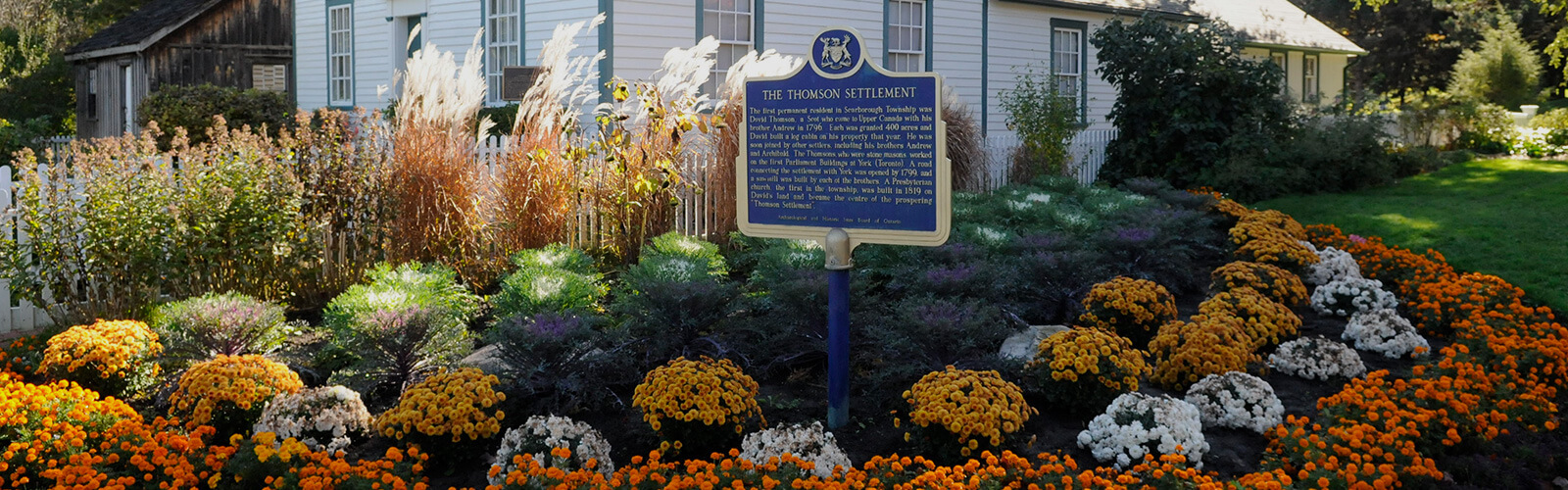 A large lush flower bed sits in front of one of the Scarborough Museum buildings. A white picket fence runs between them. A historic plaque on a post sits in front of the flower bed.