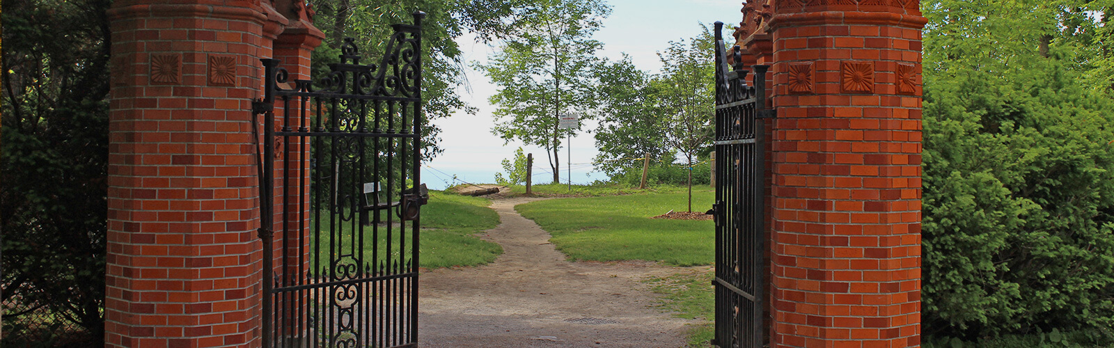 An open old looking and ornate red brick and wrought iron gate leading down a small dirt path toward a cliff edge. Lake Ontario can be seen in the background. Lush trees sit on either side of the gate.