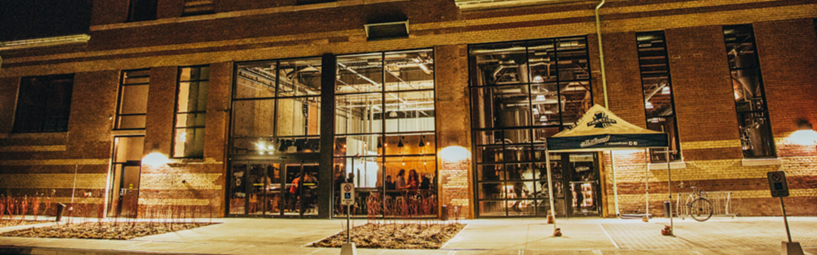 Night-time photo of large industrial looking brick building with several large windows. The interior is well lit, and inside one of the windows, a brewery tank is visible with a promotional tent sitting outside the window. Inside the other windows, a possible seating area with glimpses of people sitting and standing around.