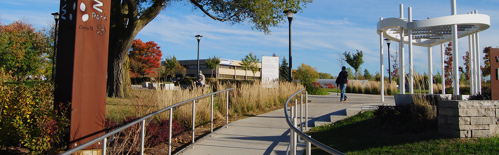 Paved ramp with metal railings on either side runs through the middle of the photo from the foreground. It leads to a paved area with a contemporary gazebo structure. Shrubs, trees and other greenery line the ramp on either side. Person can be seen walking toward the ramp in the centre.