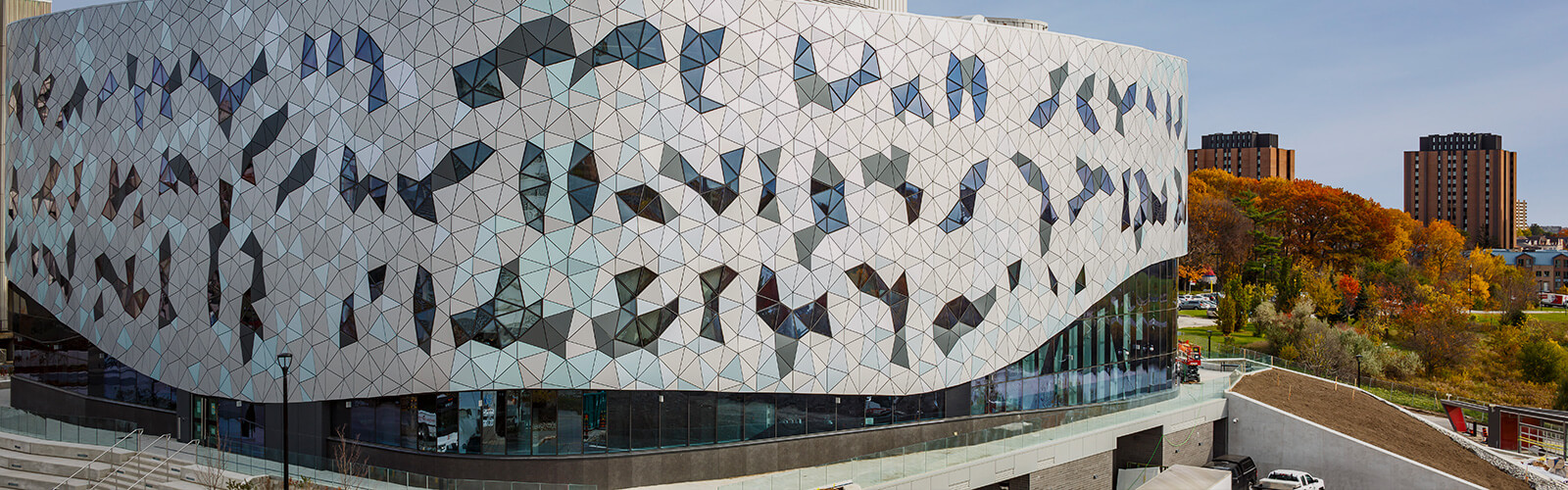 The exterior of the Bergeron Centre for Engineering Excellence-York University. It is a contemporary building with geometric glass windows stretching from one side of the building to the other. Some windows are frosted and others aren't, forming more geometric patterns within the design. The building is a low rise that takes up most of the image in the foreground. In the background are 2 brown brick apartment buildings and several fall coloured trees.