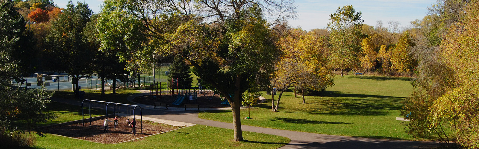 Looking down at Albion Gardens Park from a hilltop. There are children playing on the playground swingset which is surrounded by a maintained lawn, and lush autumn trees. Behind the playground is a fenced tennis court. A paved path winds through the park.